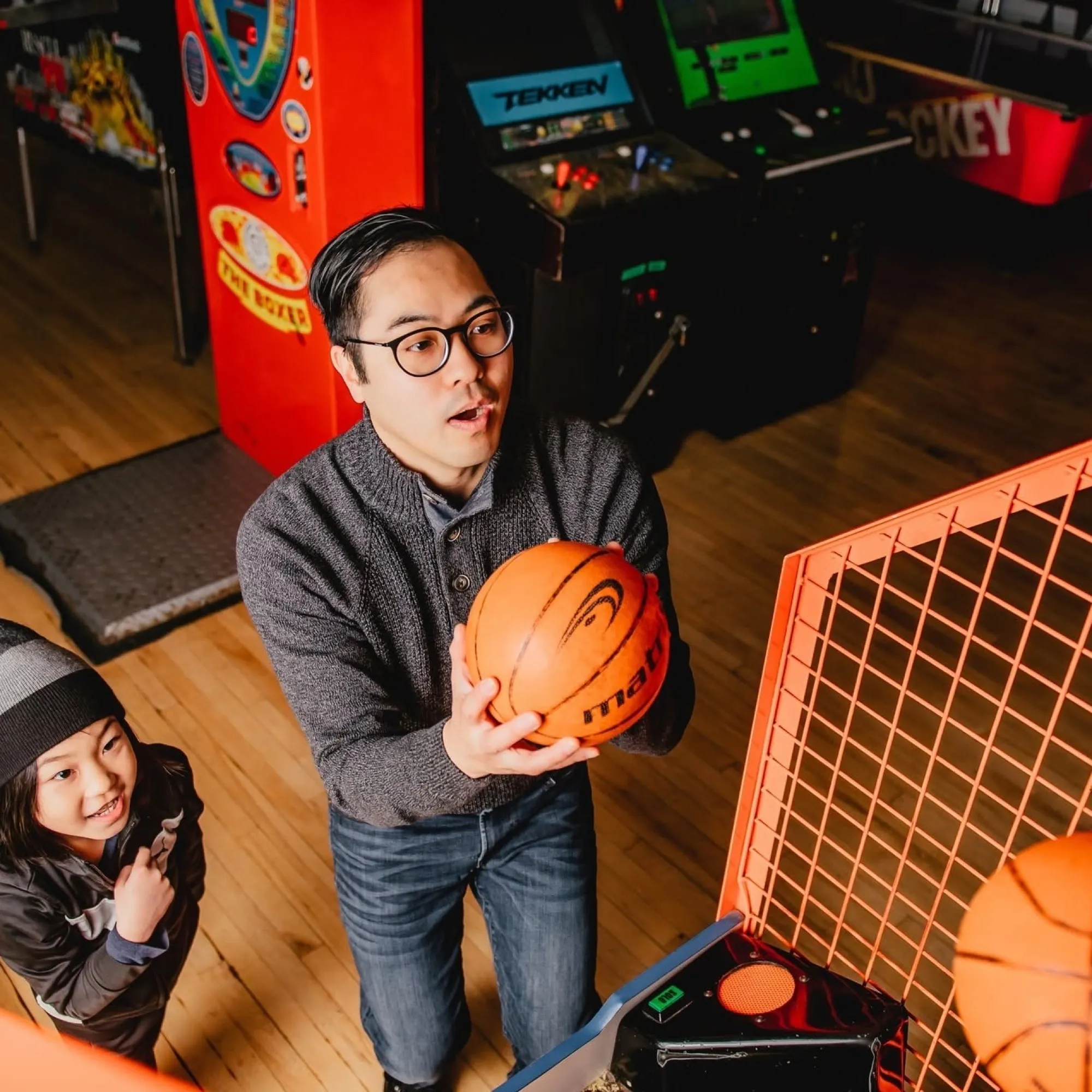 A dad lining up a shot at the arcade basketball game while a kid watches