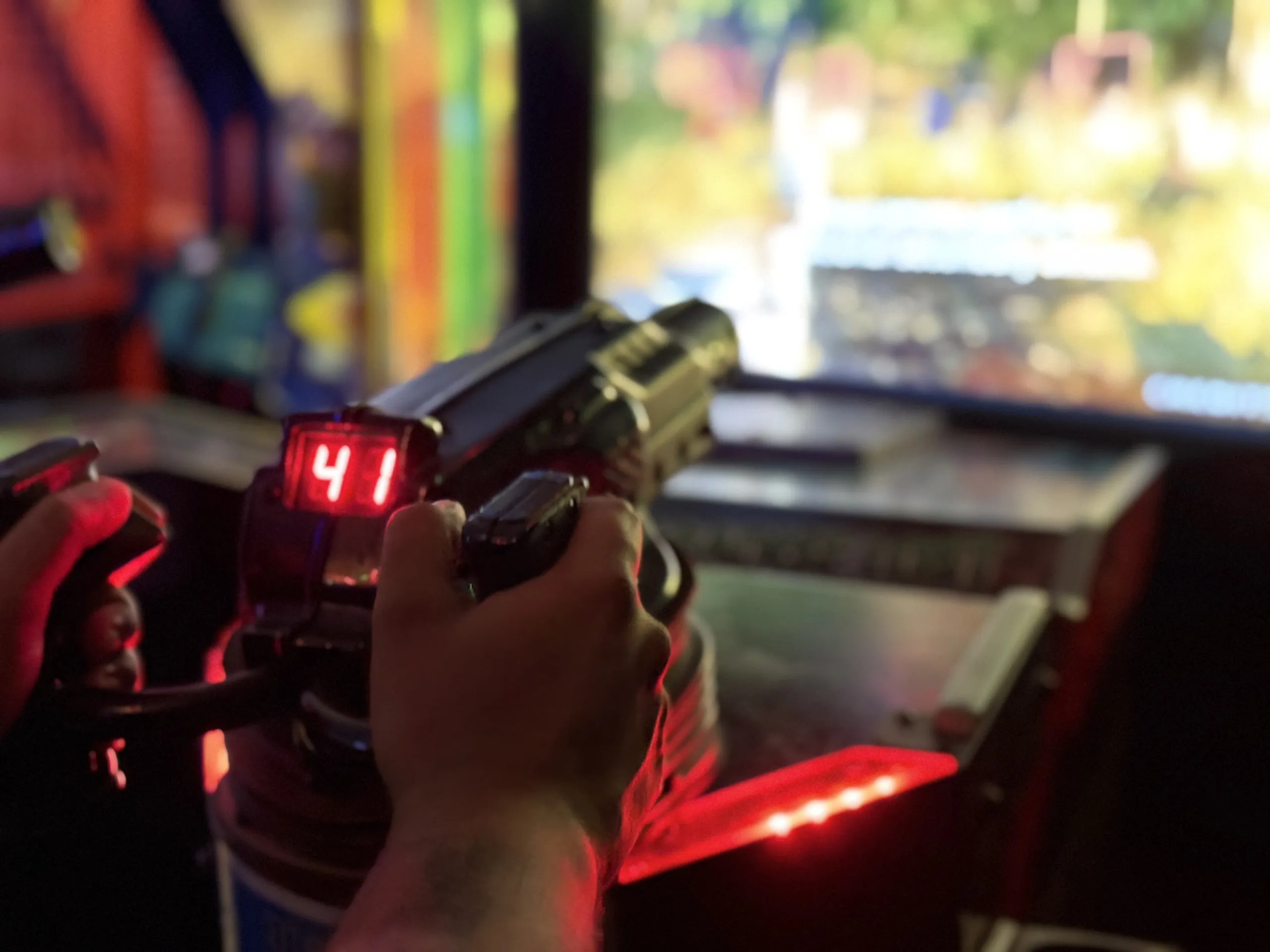 Hands gripping a light-gun controller in front of a glowing arcade shooter screen